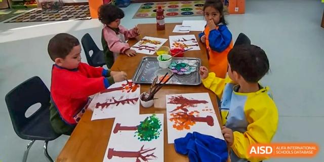 Young students in colorful smocks using paint trays to create vibrant tree artwork during an art lesson at Ecole Française Internationale de Wuhan.