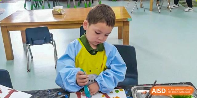 A young boy in a blue and yellow smock focuses intently on his painting project at a classroom table at Ecole Française Internationale de Wuhan.