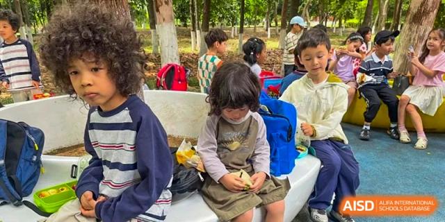Young children sit together on white benches outdoors, focused on their snacks during a school outing at Ecole Française Internationale de Wuhan.