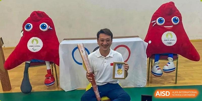 A man proudly displays an Olympic torch and a gold medal while seated in front of the Olympic rings logo at Ecole Française Internationale de Wuhan.