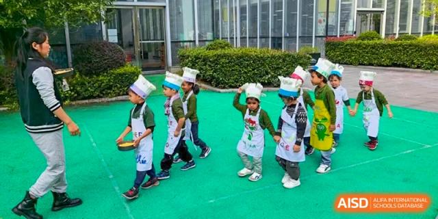 A line of young children wearing white chef hats and aprons walks across a green outdoor play area at Ecole Française Internationale de Wuhan.