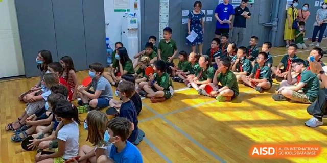 A class of students sits attentively on a wooden floor during an indoor group lesson or presentation at Ecole Française Internationale de Wuhan.