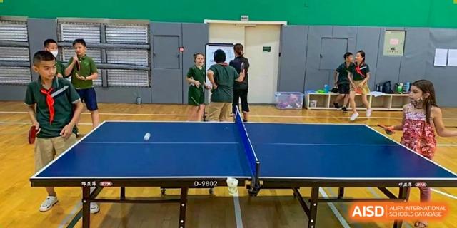 A blue ping pong table in a gymnasium with a ball in motion and students in the background at Ecole Française Internationale de Wuhan.