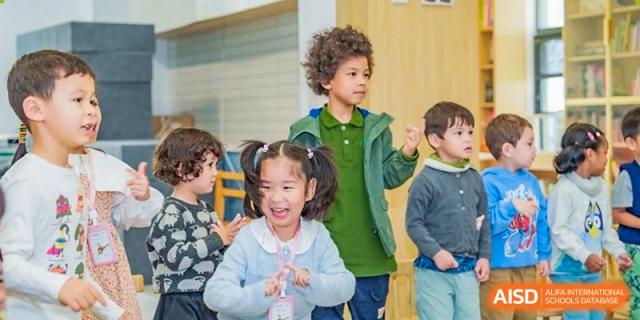 A group of young children laughing and playing together indoors, with one girl in a blue sweater smiling at Ecole Française Internationale de Wuhan.