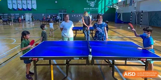 Two adults and a student standing by a professional blue table tennis table inside a large green-walled gym at Ecole Française Internationale de Wuhan.