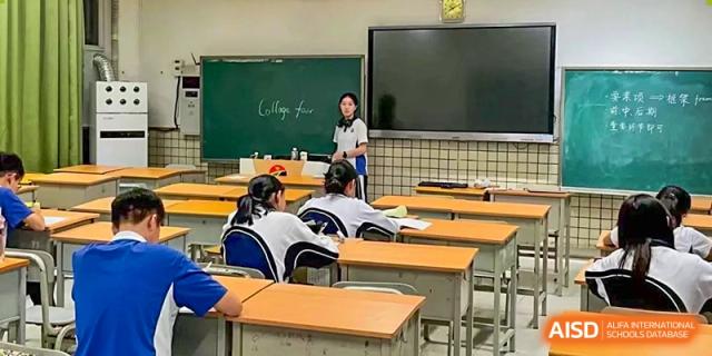 A classroom view from the back showing students at wooden desks facing a teacher standing at a green chalkboard at Shenzhen Senior High School International Division.