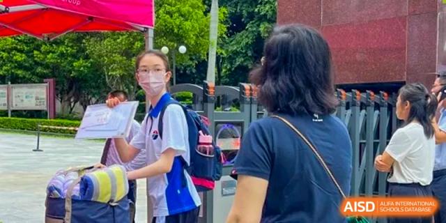A female student wearing a face mask and a white school polo shirt walking through an outdoor campus gate at Shenzhen Senior High School International Division.