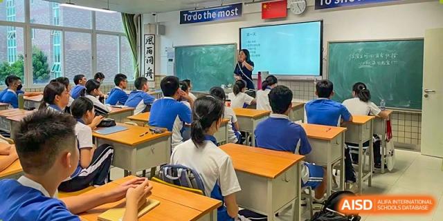 A teacher leading a lesson in a bright classroom with students seated at light wood desks facing a screen at Shenzhen Senior High School International Division.