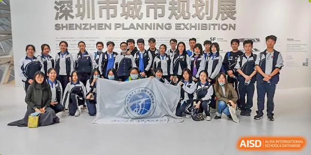 A large group of students posing with a white school flag in front of a "Shenzhen Planning Exhibition" sign at Shenzhen Senior High School International Division.