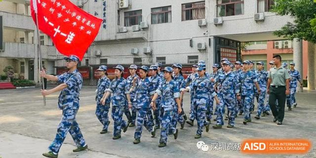 Students in blue camouflage military training uniforms march in formation across a paved courtyard at Shenzhen Senior High School International Division.