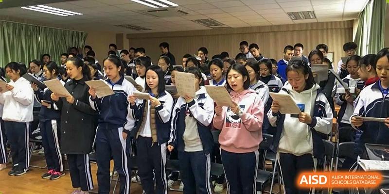 Students in tracksuits stand together indoors holding papers and singing or reciting during a rehearsal at Shenzhen Senior High School International Division.