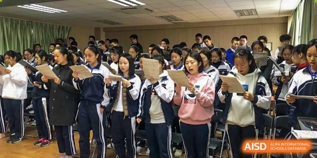 Students in tracksuits stand together indoors holding papers and singing or reciting during a rehearsal at Shenzhen Senior High School International Division.