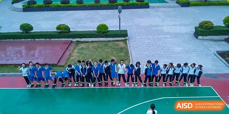 A line of students in blue uniforms stands on a green outdoor court, bowing together in a synchronized gesture at Shenzhen Senior High School International Division.