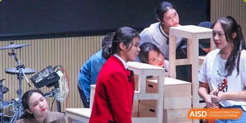 Students in formal red and blue blazers work together to assemble white wooden crates on a stage for a performance at Shenzhen Senior High School International Division.
