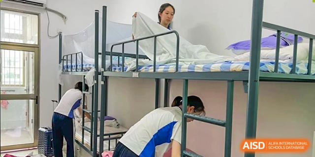 A student in a white and blue uniform making a bed in a dormitory room with bunk beds at Shenzhen Senior High School International Division.