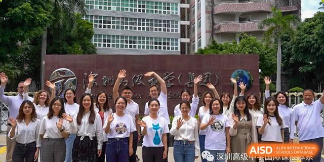 Faculty and staff members smiling and waving in front of the campus entrance gate at Shenzhen Senior High School International Division.
