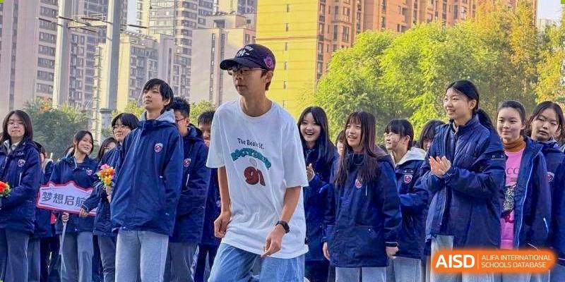 A student in a white t-shirt and baseball cap stands outdoors with classmates in blue jackets at Wuxi Foreign Language School International Curriculum Center.