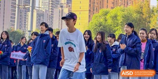 A student in a white t-shirt and baseball cap stands outdoors with classmates in blue jackets at Wuxi Foreign Language School International Curriculum Center.