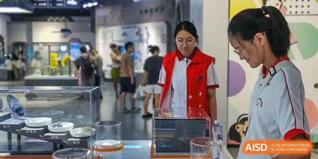 A student volunteer in a red vest stands by a glass display case featuring scientific exhibits and educational posters at Wenzhou Foreign Language School.