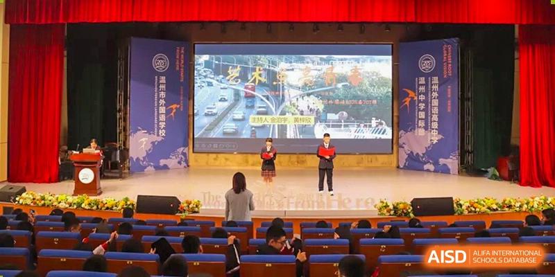 Two students presenting on a stage in front of a large screen and a seated audience in an auditorium at Wenzhou Foreign Language School.