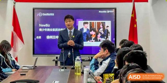 A male speaker in a suit stands before a digital screen giving an educational presentation to a group of students at XuZhou No.3 Middle School.