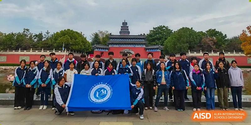 A large group of students in blue tracksuits poses with a blue school flag in front of a traditional Chinese pagoda at XuZhou No.3 Middle School.