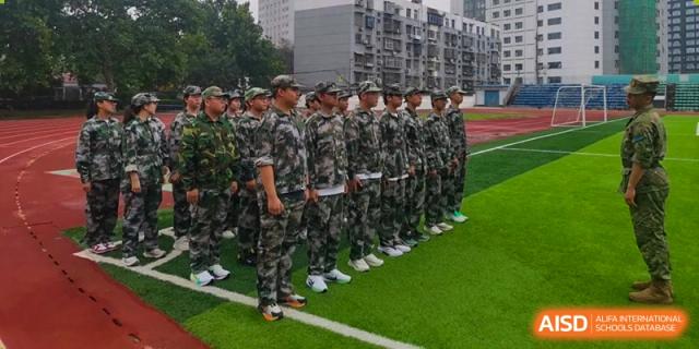 A large group of students in matching camouflage uniforms stands in neat rows on a green sports field at XuZhou No.3 Middle School.