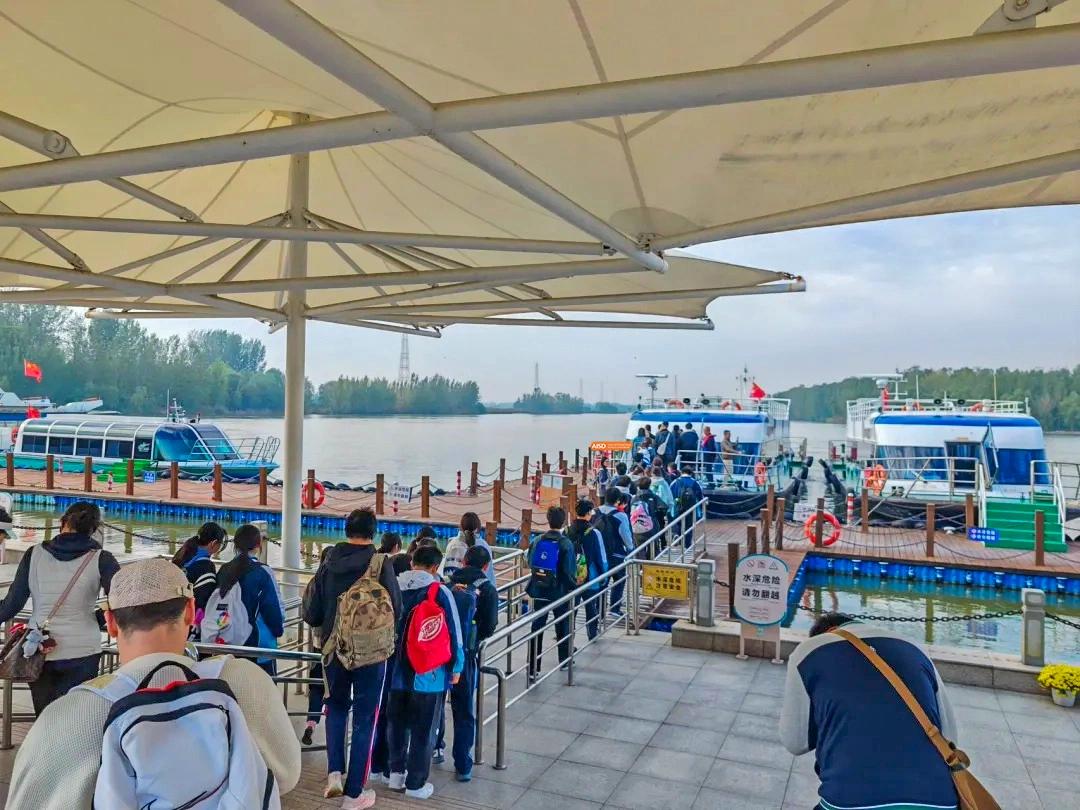 A large group of students gathers at a waterfront pier to board a ferry during a school-organized field trip at XuZhou No.3 Middle School.
