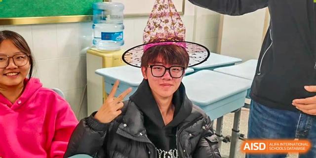 A smiling student wearing glasses and a festive pointed hat gives a peace sign while sitting in a classroom at XuZhou No.3 Middle School.