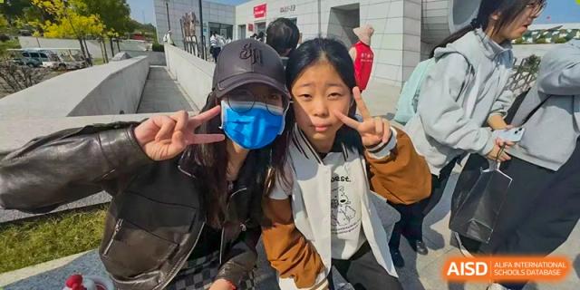Two female students smile for a close-up photo, one wearing a black baseball cap and a blue face mask at XuZhou No.3 Middle School.