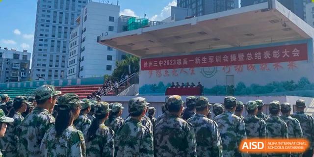 A large group of students in matching camouflage military attire stands in formation during an outdoor ceremony at XuZhou No.3 Middle School.