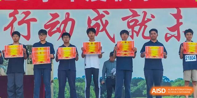 Three students stand on a stage proudly holding up official red and gold award certificates for their achievements at XuZhou No.3 Middle School.