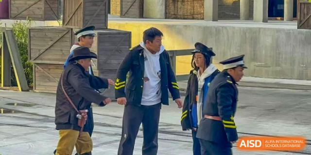 A teacher in a dark coat interacts with students outdoors near the school buildings during a break between classes at XuZhou No.3 Middle School.