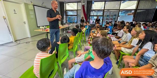 A man in a black shirt stands before a group of students seated in green chairs in a bright modern classroom at École Française Internationale de Canton(EFIC)