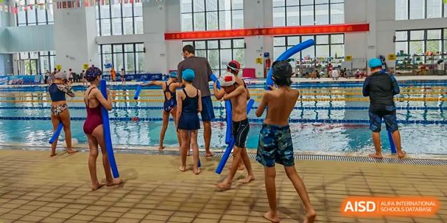 Students in swimwear stand at the edge of an indoor pool while an instructor assists them with blue foam noodles at École Française Internationale de Canton(EFIC)