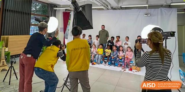 A photographer in a yellow vest sets up a group photo of young students sitting on benches in a studio at École Française Internationale de Canton(EFIC)