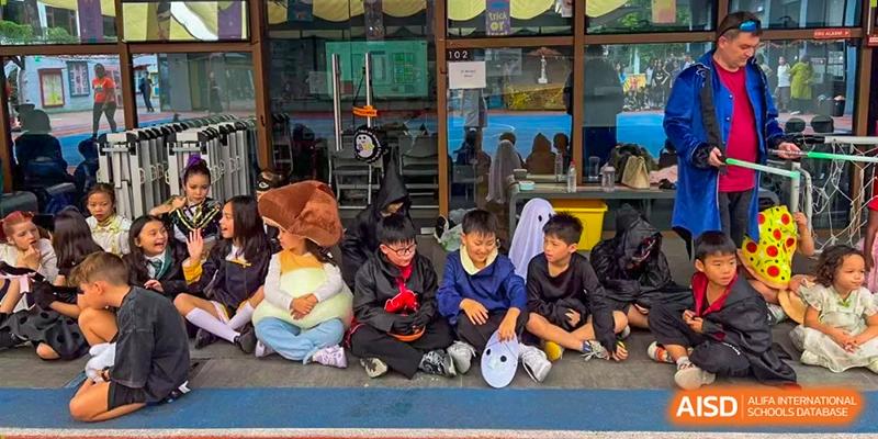 A group of children dressed in various Halloween costumes sit together on a blue mat in a classroom setting at École Française Internationale de Canton(EFIC)