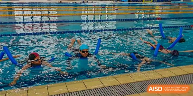 A young student wearing goggles and a blue swim cap practices swimming with a blue foam noodle in a lap pool at École Française Internationale de Canton(EFIC)