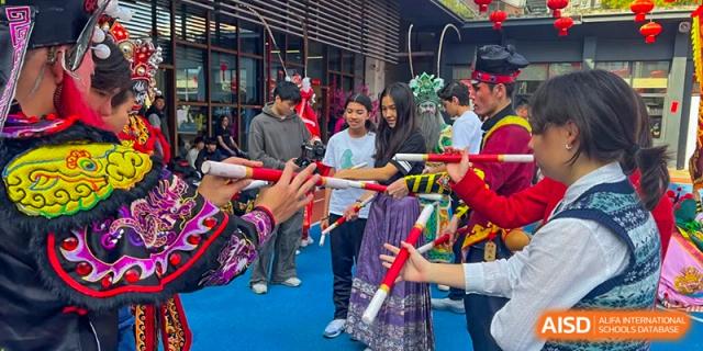 A group of students engage in a traditional folk dance using long decorated poles on an outdoor court at École Française Internationale de Canton(EFIC)