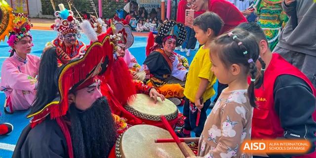 A performer in a traditional red costume plays a large drum while a student watches closely at École Française Internationale de Canton(EFIC)