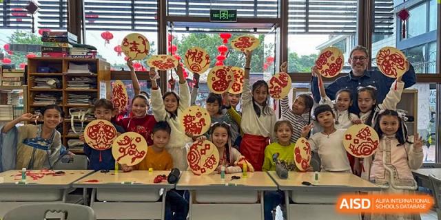 A smiling group of children hold up round decorative crafts with red designs in a classroom at École Française Internationale de Canton(EFIC)