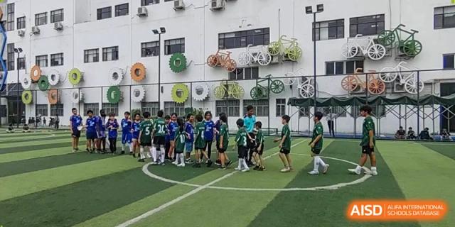 Children in green and blue jerseys gather on an outdoor turf soccer field for a game, with bicycle art decorating the building behind them at École Française Internationale de Canton(EFIC).