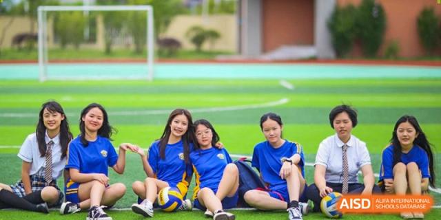 Three female students in blue athletic wear sit and smile together on the green sports field at Beijing International Bilingual Academy Guiyang.