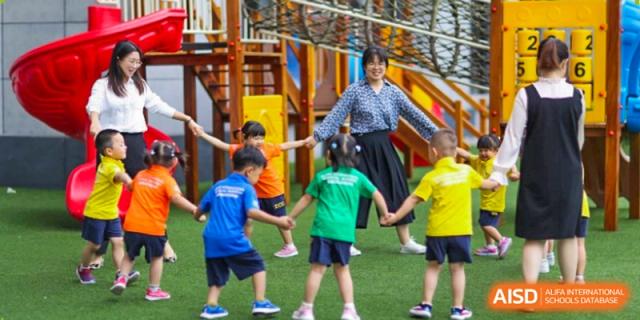 A teacher in a patterned blouse plays a circle game with young children on the outdoor playground at Beijing International Bilingual Academy Guiyang.