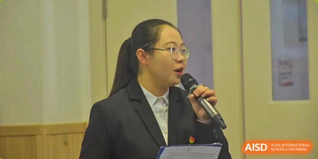 A female student in a formal suit speaks into a microphone during a school presentation or assembly at Beijing International Bilingual Academy Guiyang.