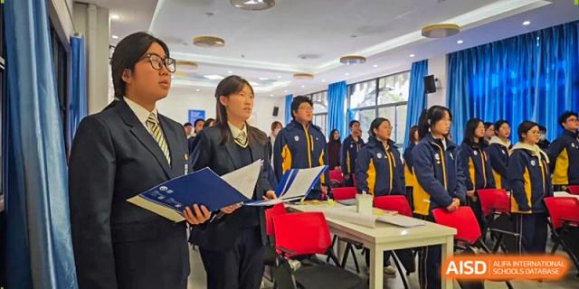 A student leader stands at the front of a classroom holding a blue folder during a school meeting at Beijing International Bilingual Academy Guiyang.