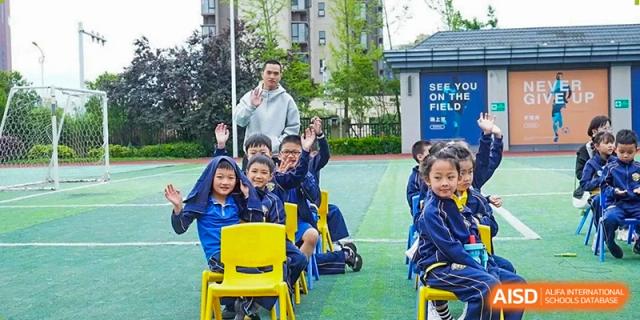 Students sit in yellow chairs on the field and wave at the camera during an outdoor school assembly at Beijing International Bilingual Academy Guiyang.