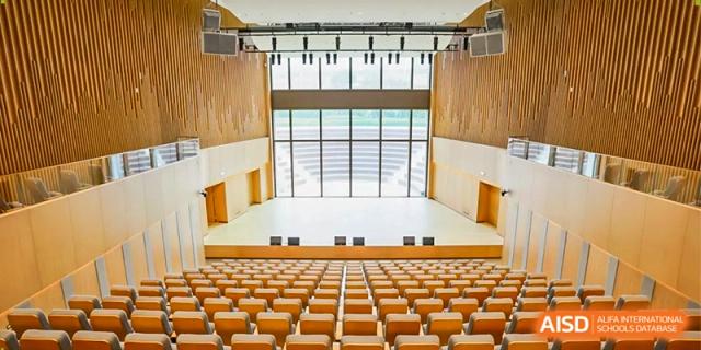 A high-angle view of a large, modern school auditorium with tiered wooden seating facing a bright stage at Benenden Bilingual School Guangzhou
