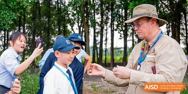 A group of students wearing school caps participate in an outdoor nature lesson in a wooded area at Benenden Bilingual School Guangzhou