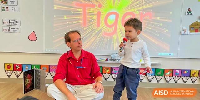 A teacher in a red shirt interacts with a young student holding a microphone in front of a colorful digital screen at Benenden Bilingual School Guangzhou